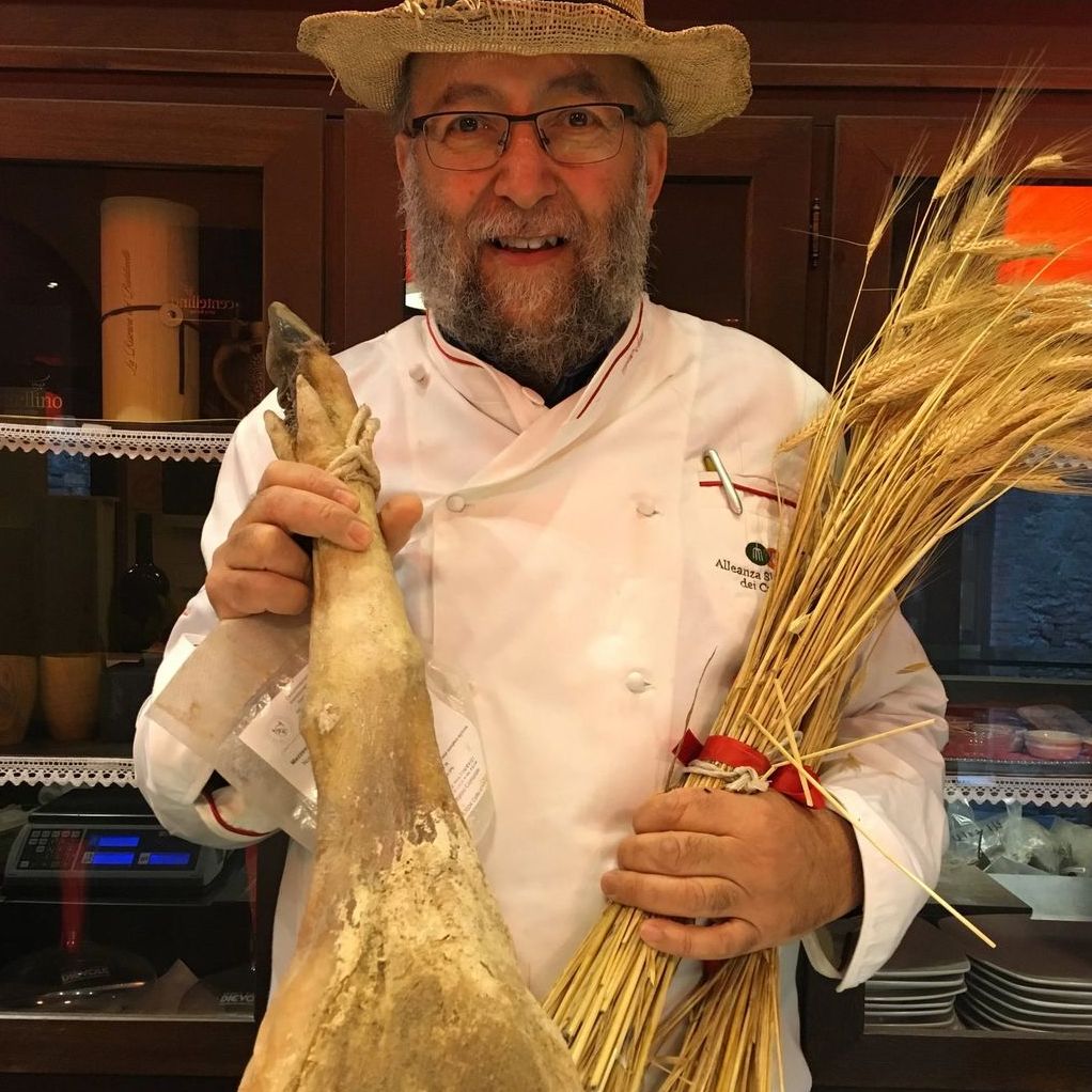 Angelo Senes in giacca da cuoco e cappello di paglia, con un prosciutto di Cinta e un mazzo di spighe